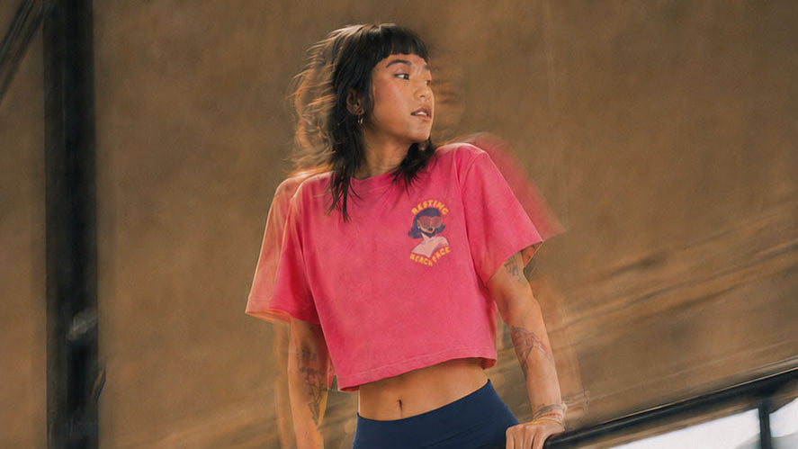 A woman in a pink cropped t-shirt and navy blue athletic shorts performing a dip on a parallel bar in an industrial loft setting.