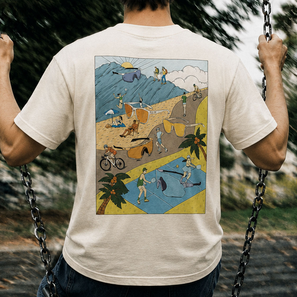 A man viewed from behind wearing a cream-colored t-shirt with a colorful graphic print on the back, hanging on a swing in an outdoor park setting.
