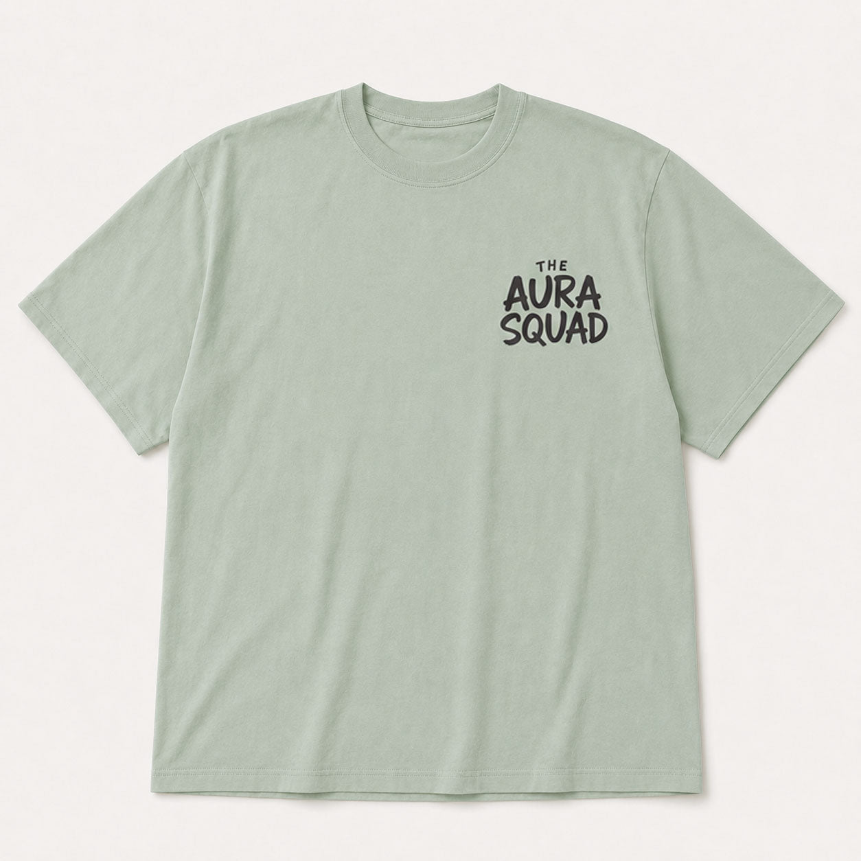 A light sage green crewneck t-shirt with 'THE AURA SQUAD' printed in black on the left chest, displayed flat against a light off-white background.