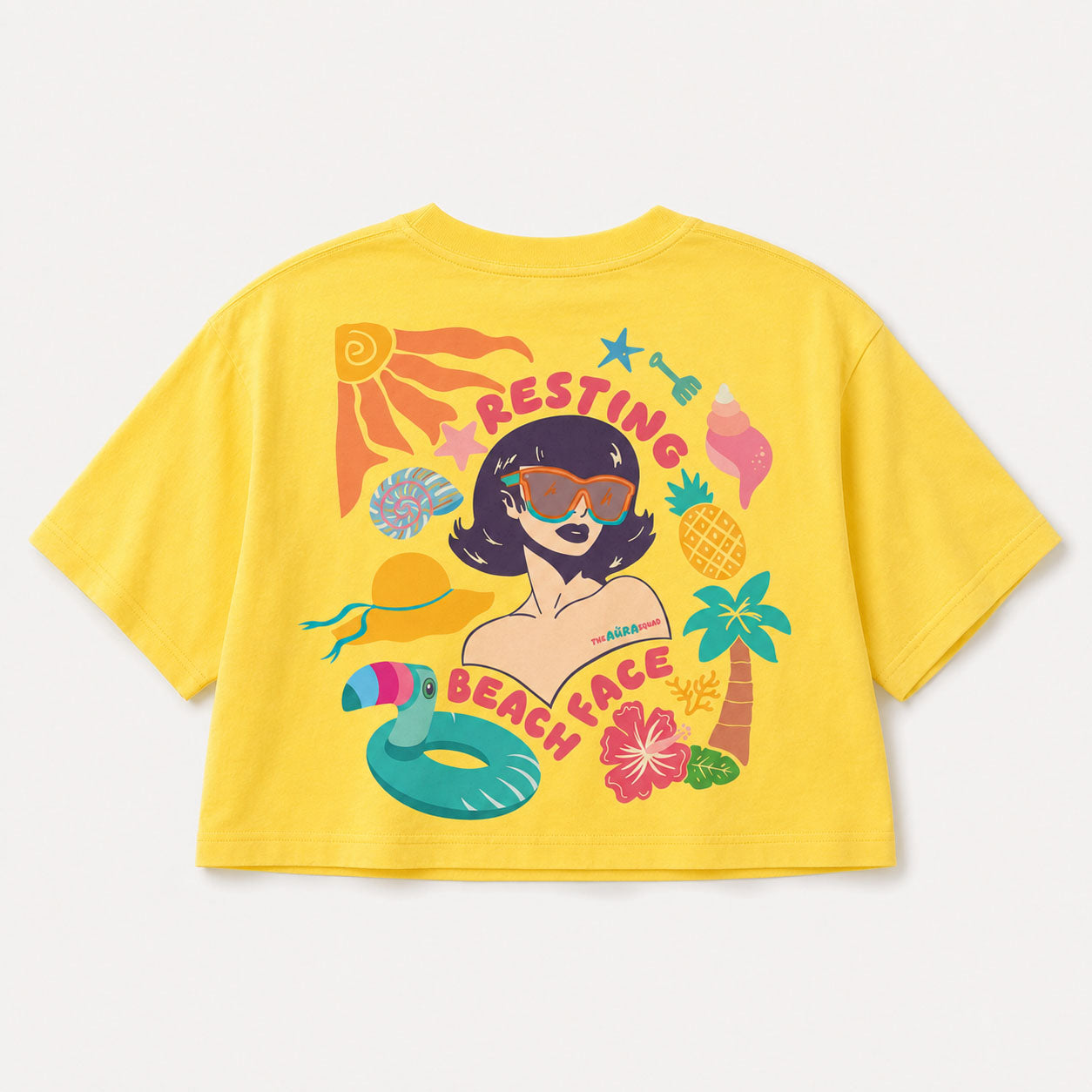 A bright yellow short-sleeve crop top t-shirt featuring a colorful retro-style graphic of a woman with sunglasses surrounded by beach-themed illustrations like a palm tree, pineapple, and pool float, with the text 'RESTING BEACH FACE'.