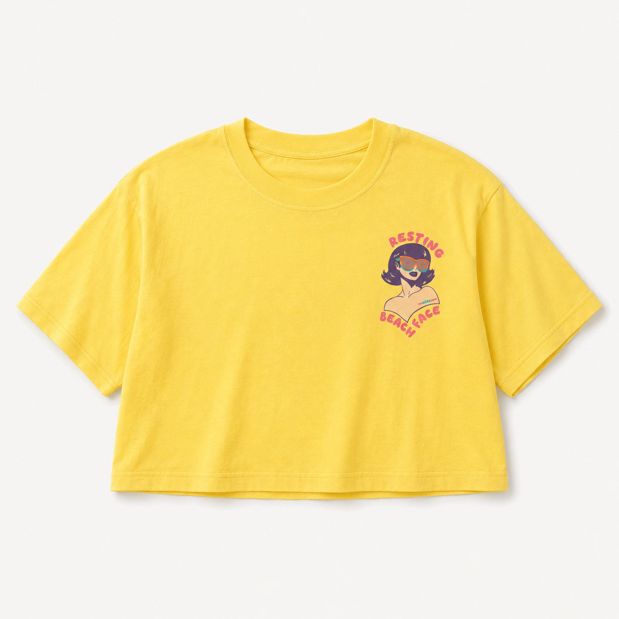 A bright yellow cropped short-sleeve t-shirt with a small graphic print on the left chest showing a retro-style woman with pink hair and the text 'RESTING BEACH FACE' in pink, displayed on a clean off-white background.