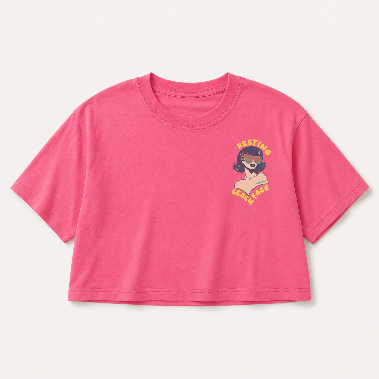 A bright pink short-sleeve crop top t-shirt with a small graphic on the left chest showing a woman with sunglasses and the text 'RESTING BEACH FACE' in yellow, displayed on a light off-white background.