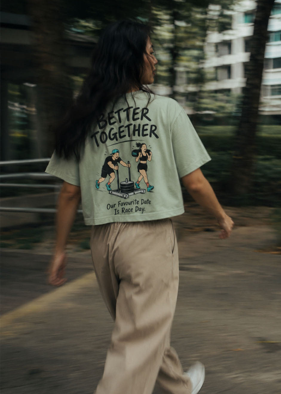 A woman walking outdoors on a path, viewed from behind, wearing a light sage green graphic t-shirt with the text 'BETTER TOGETHER' and a cartoon illustration on the back, paired with beige trousers and white sneakers.