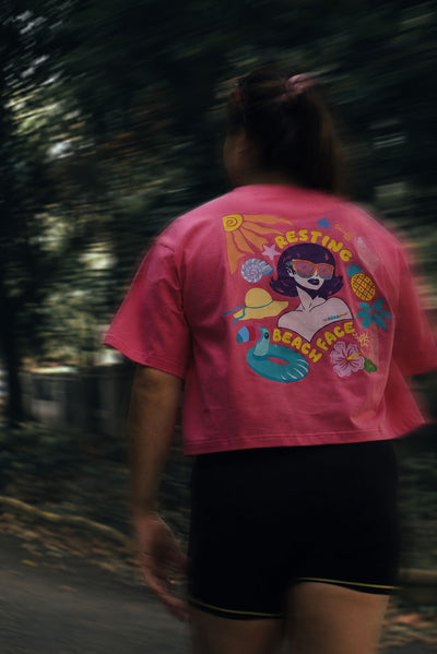 A person viewed from behind wearing a bright pink cropped T-shirt with a colorful retro beach-themed graphic on the back, paired with black shorts, set against a blurred dark green outdoor background.