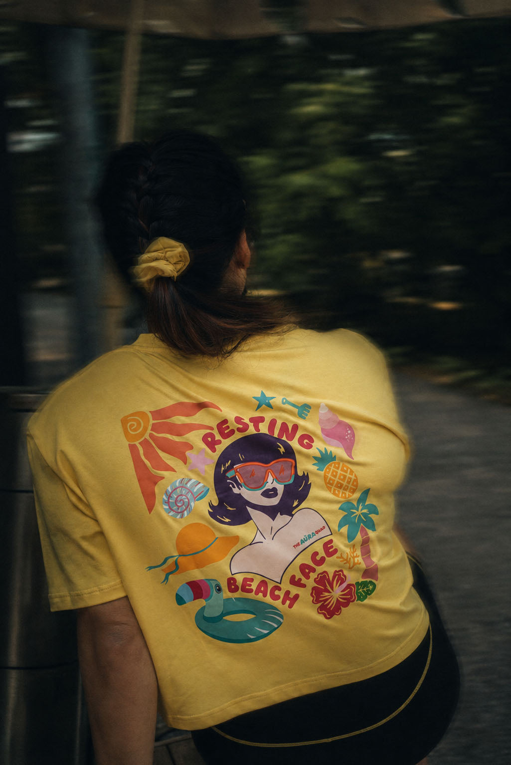 A person viewed from behind wearing a bright yellow graphic T-shirt with a colorful retro beach-themed print and the words 'RESTING BEACH' on the back, photographed outdoors with a blurred green background.