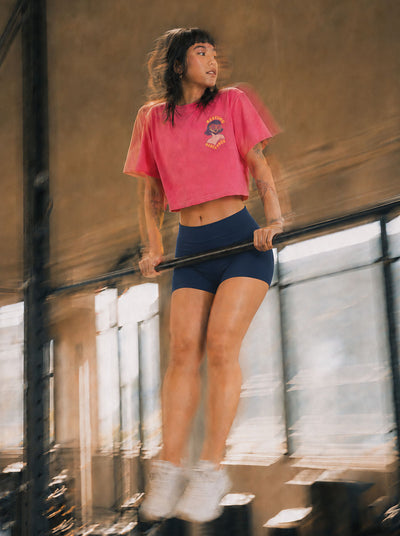 A woman in a pink cropped t-shirt and navy blue athletic shorts performing a dip on a parallel bar in an industrial loft setting.