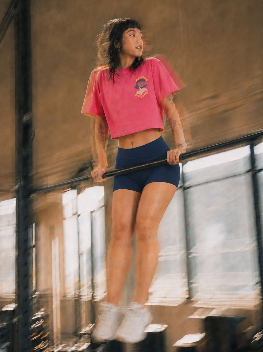 A woman in a pink cropped t-shirt and navy blue athletic shorts performing a dip on a parallel bar in an industrial loft setting.