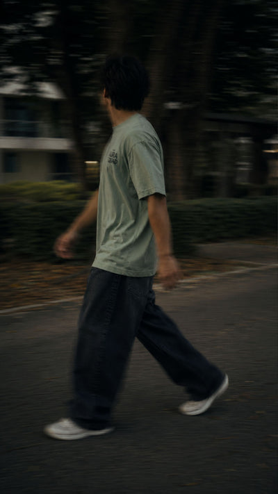A person walking outdoors in a light sage green t-shirt, dark wide-leg jeans, and white sneakers on a paved path with blurred trees in the background.