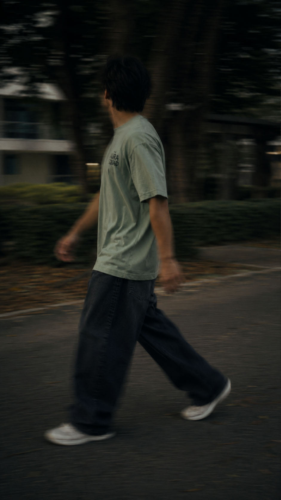 A person walking outdoors in a light sage green t-shirt, dark wide-leg jeans, and white sneakers on a paved path with blurred trees in the background.