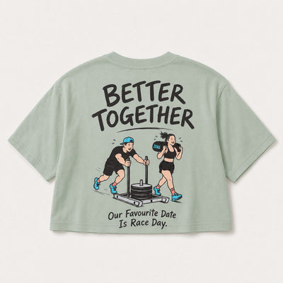 A sage green short-sleeve t-shirt shown from the back, featuring a cartoon graphic of a man pushing a weight stack and a woman boxing, with the text 'BETTER TOGETHER' and 'Our Favourite Date Is Race Day.'.