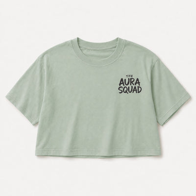 A light sage green cropped short-sleeve t-shirt with 'THE AURA SQUAD' printed in black on the left chest, displayed flat against a light gray background.