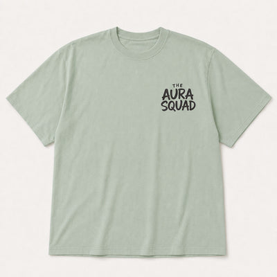 A light sage green crewneck t-shirt with 'THE AURA SQUAD' printed in black on the left chest, displayed flat against a light off-white background.