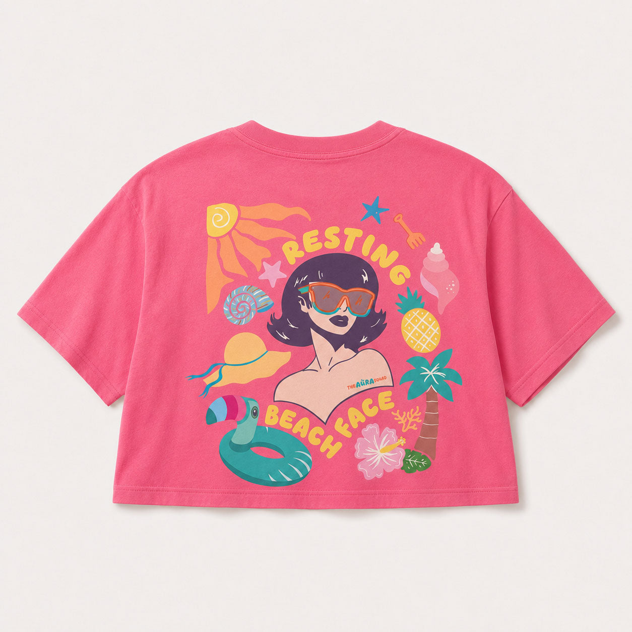 A bright pink short-sleeve graphic T-shirt shown flat against a light beige background. The back of the shirt features a colorful cartoon beach-themed design with the words 'RESTING BEACH FACE,' a woman with sunglasses, a flamingo float, palm tree, pineapple, and other summer motifs.