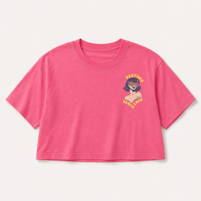 A bright pink short-sleeve crop top t-shirt with a small graphic on the left chest showing a woman with sunglasses and the text 'RESTING BEACH FACE' in yellow, displayed on a light off-white background.