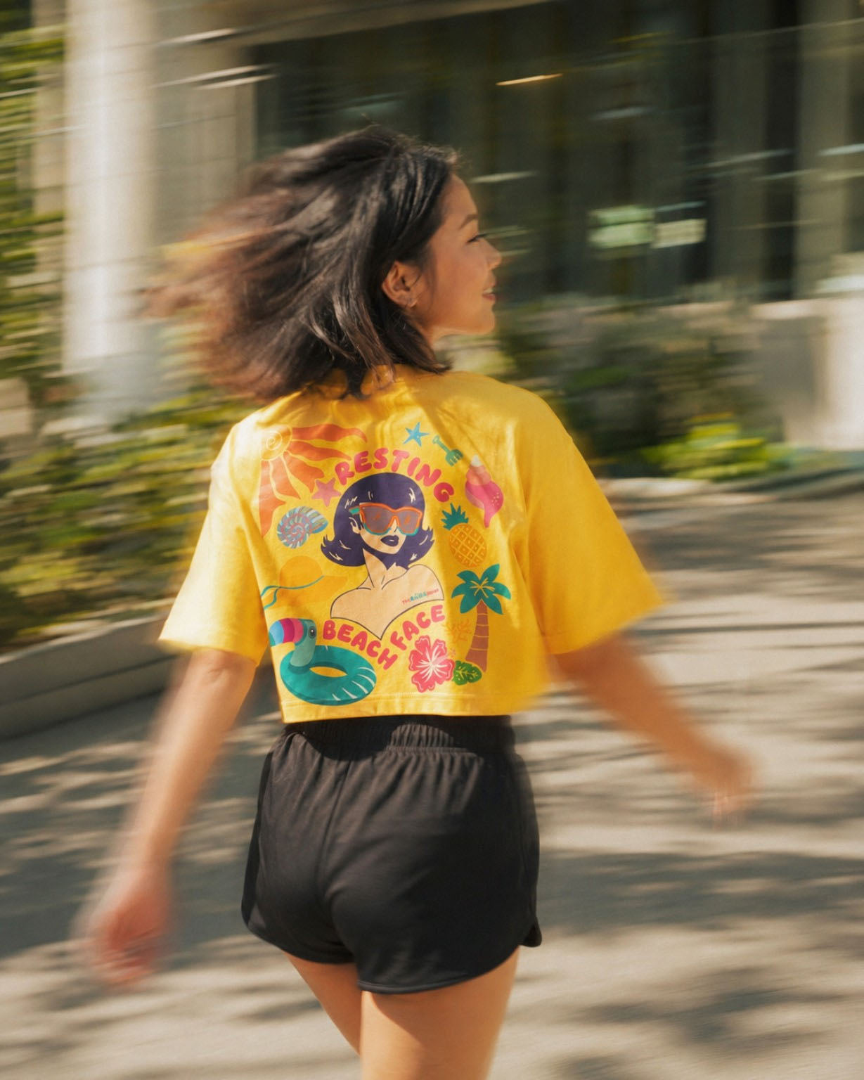 A woman wearing a bright yellow graphic t-shirt and black shorts, captured from behind as she walks outdoors. The back of the t-shirt features a colorful retro-style illustration of a woman at the beach with the text 'FISHING FOR A BEACH GIRL'.
